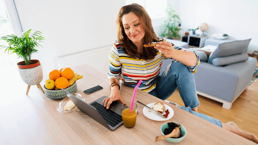 benefícios e vantagens de participar de programas de afiliados blog saia da crise Milla Ribeiro mulher ruiva sentada à mesa da sala de estar trabalhando no notebook enquanto faz um lanche